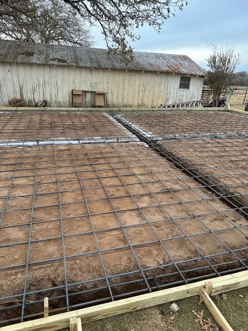 Rebar grid tied across the slab pad inside wood forms, an old wood barn beyond.