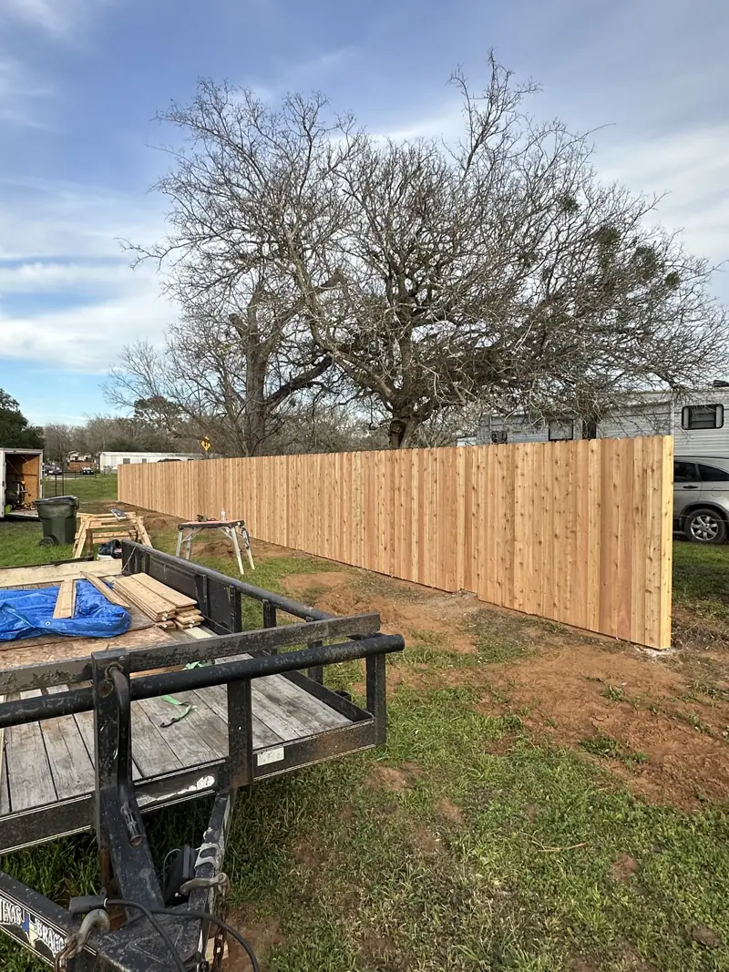 New cedar privacy fence along a property line, with a work trailer, sawhorses and spare pickets in front.