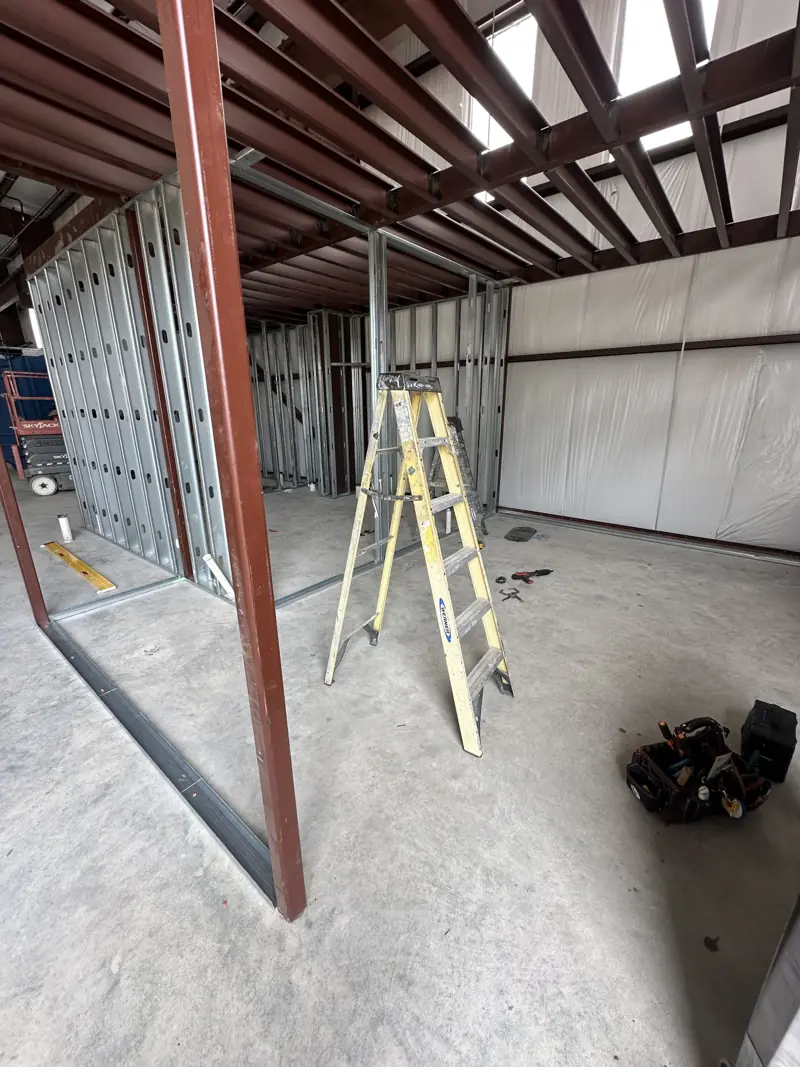 Galvanized metal stud walls under red steel mezzanine joists, with a stepladder and tool bag on the slab.