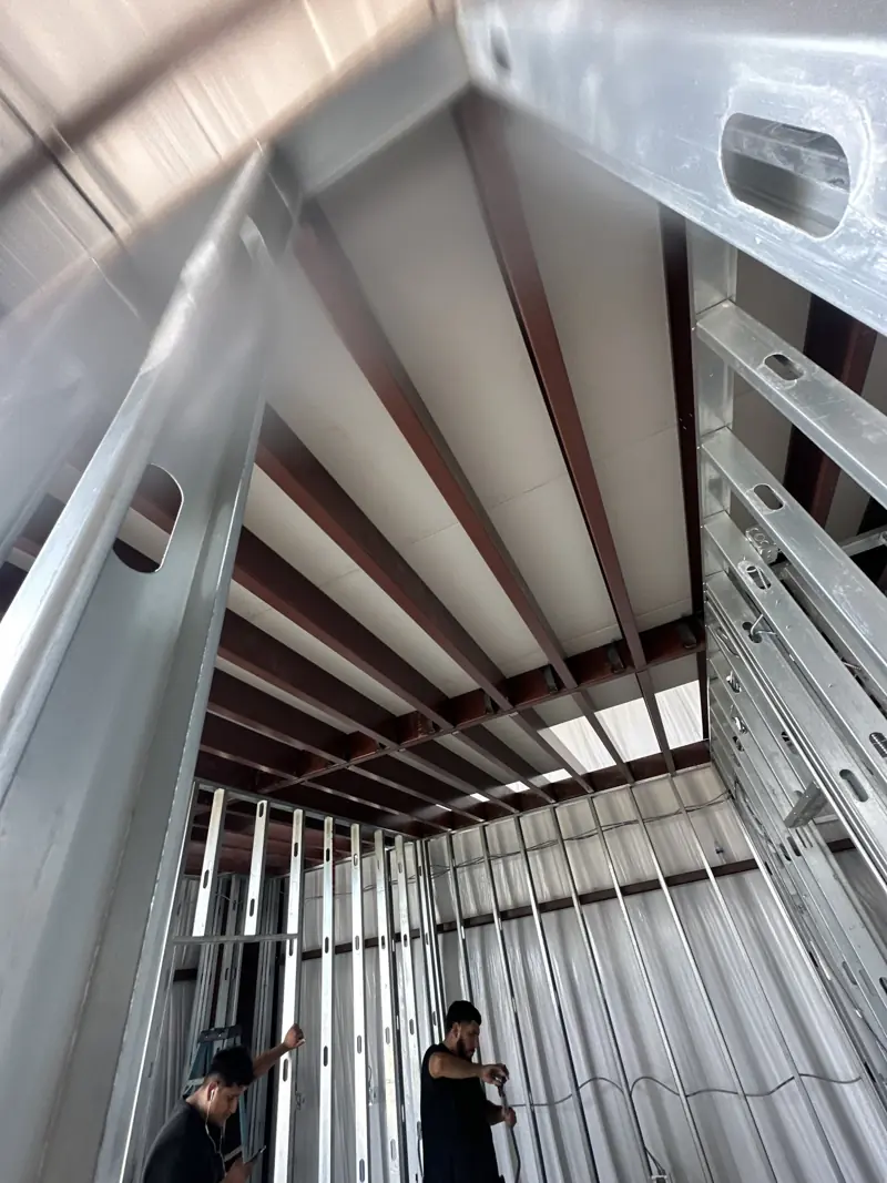 Looking up past metal studs at steel joists and board decking above, two workers framing walls below.