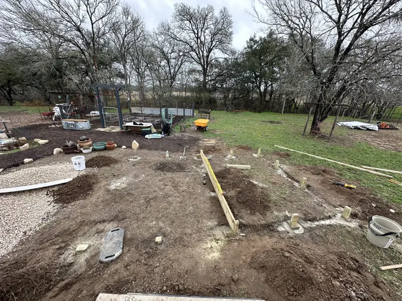 Cleared dirt pad with treated wood posts set in concrete footings, a leveling board and piles of dug soil.
