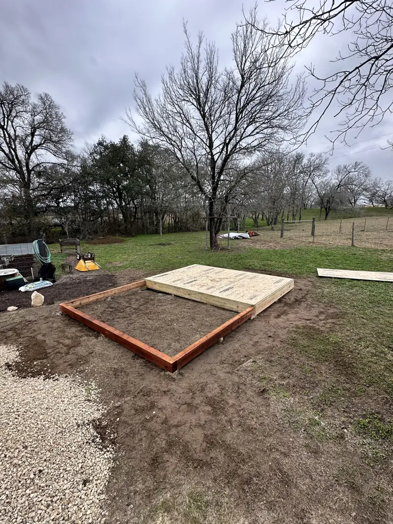 Shed floor deck sheathed in subfloor panels on treated framing, beside a red-stained timber base frame.