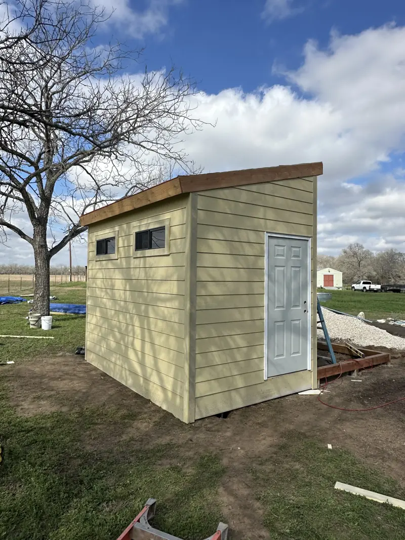 Shed with tan lap siding, two small windows, a six-panel door and a sloped roof edge, next to a gravel pile.