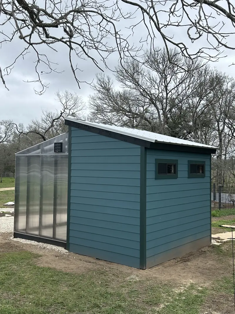 Blue lap-sided shed with two small windows, dark trim and a metal roof, with the greenhouse at left.