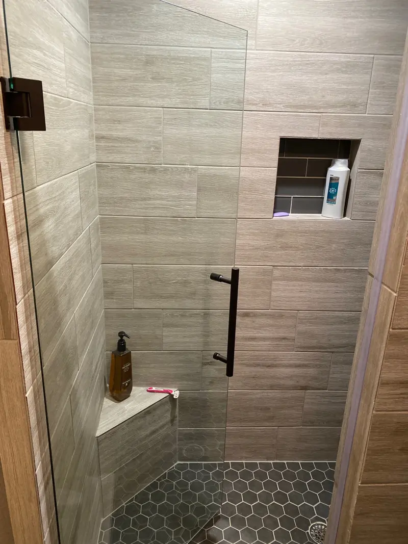 Walk-in shower with wood-look plank tile, a niche, a corner bench, a black hex tile floor and a glass door.