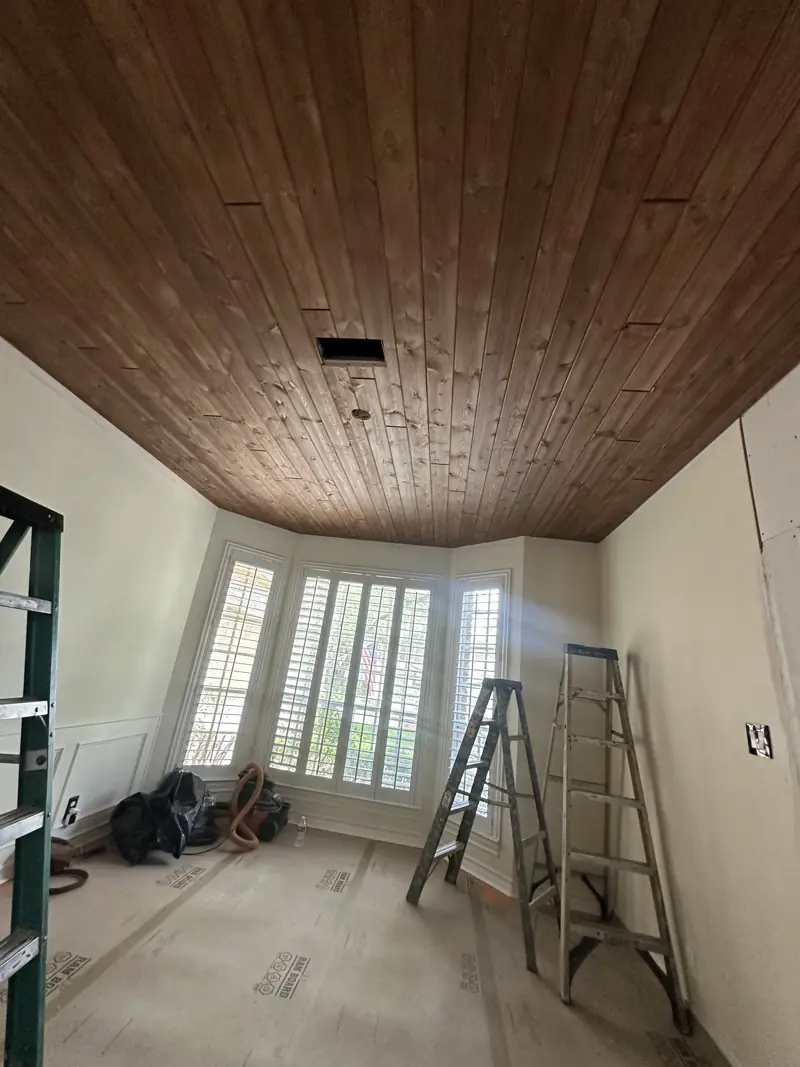 Stained wood plank ceiling over the room, with a bay window, plantation shutters and two ladders below.