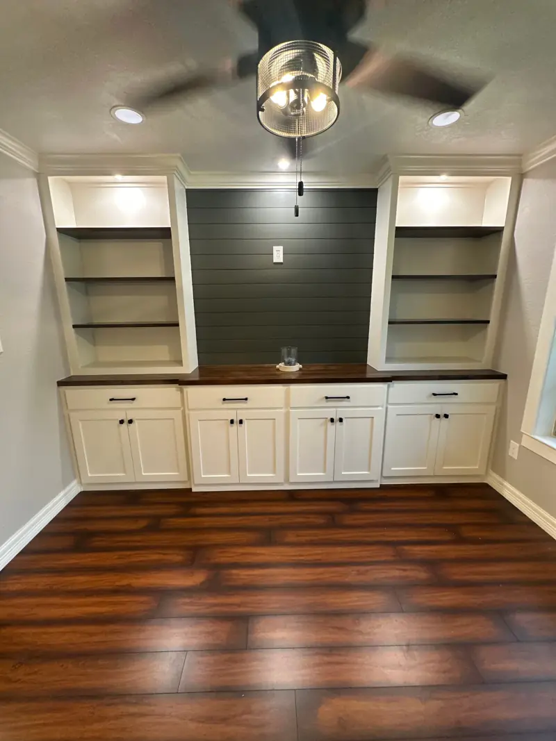 Office wall with built-in white cabinets and lit open shelves on each side of a dark shiplap wall, over a wood-look floor.