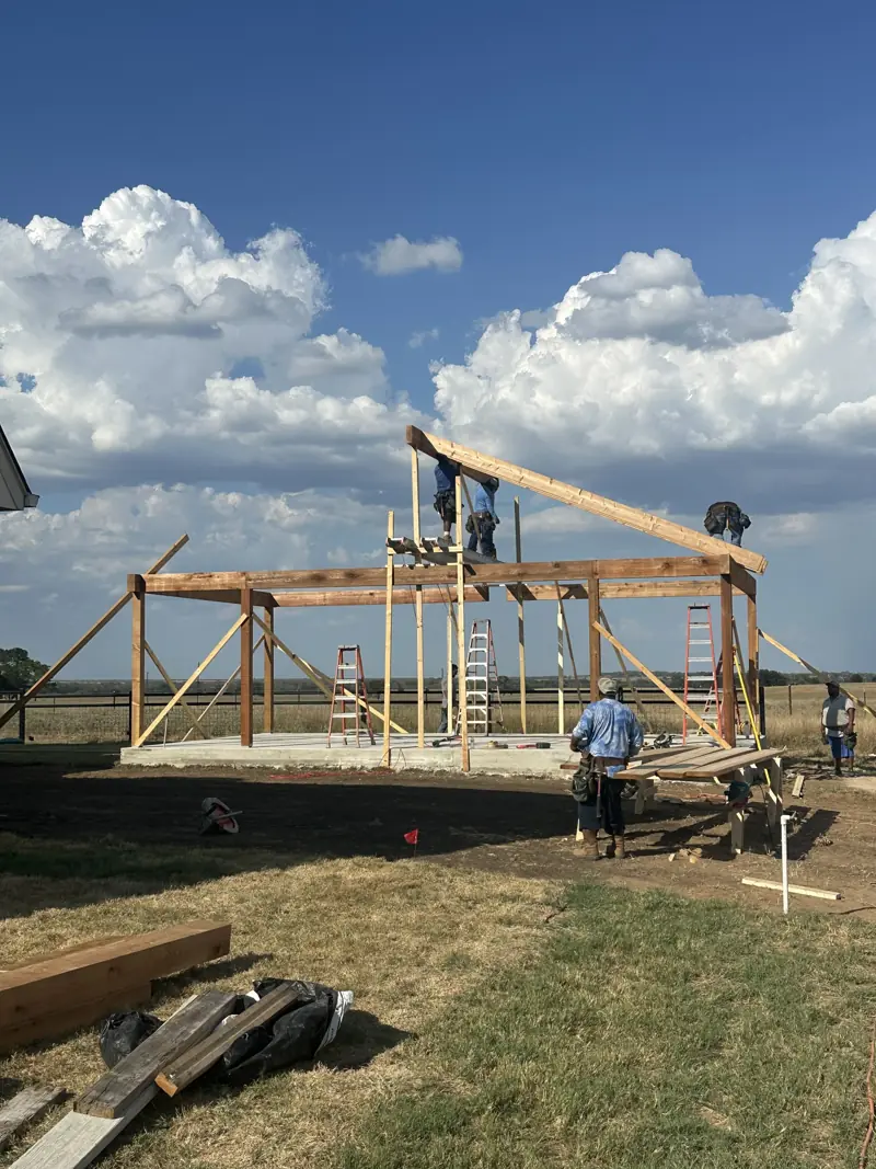 Crew on ladders and scaffold setting a sloped rafter on a timber post-and-beam frame over a new concrete slab.