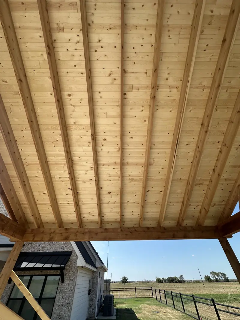 Knotty pine plank ceiling on exposed rafters over a timber beam, with a brick house and fenced pasture beyond.