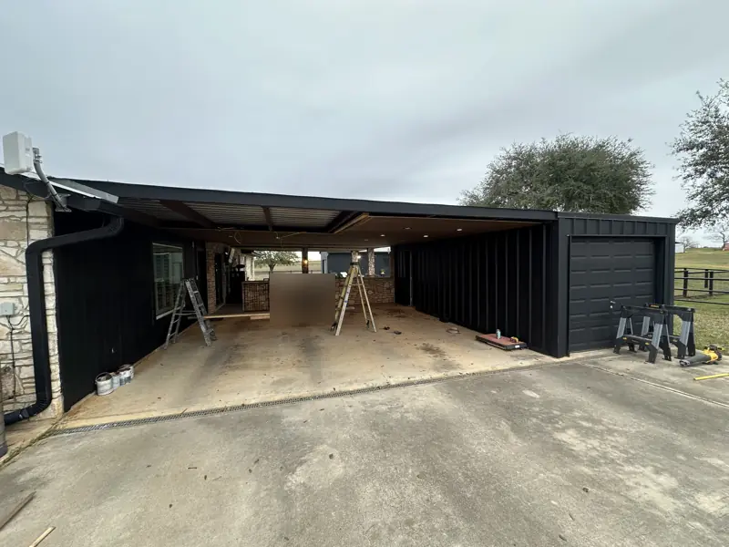 Open breezeway under a black-trimmed patio roof with the ceiling partly removed, ladders and sawhorses on the slab.