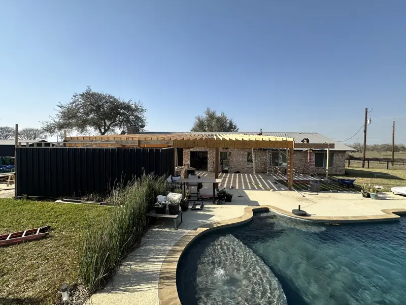 New wood roof framing over the patio along the stone house, seen across the pool on a clear day.