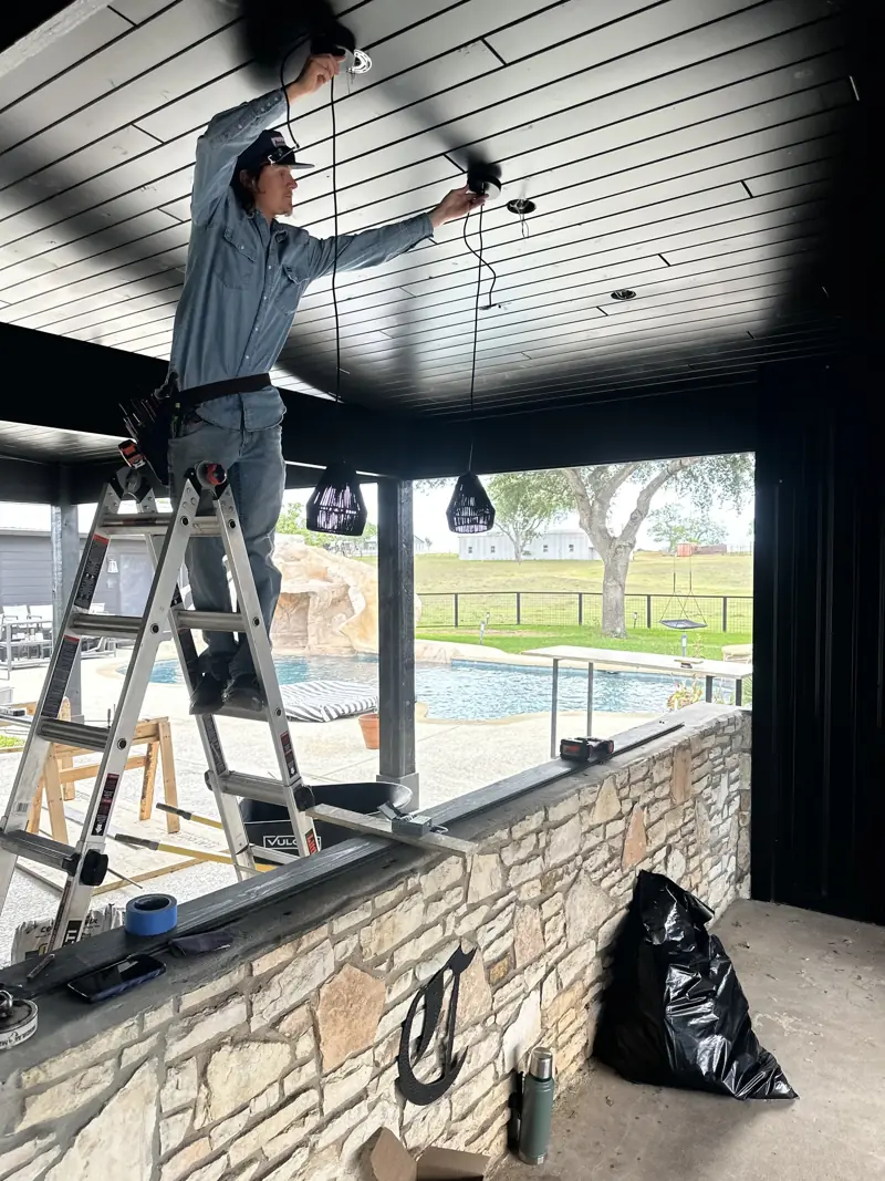 Worker on a ladder hanging black pendant lights from the new black plank ceiling above a stone bar wall.