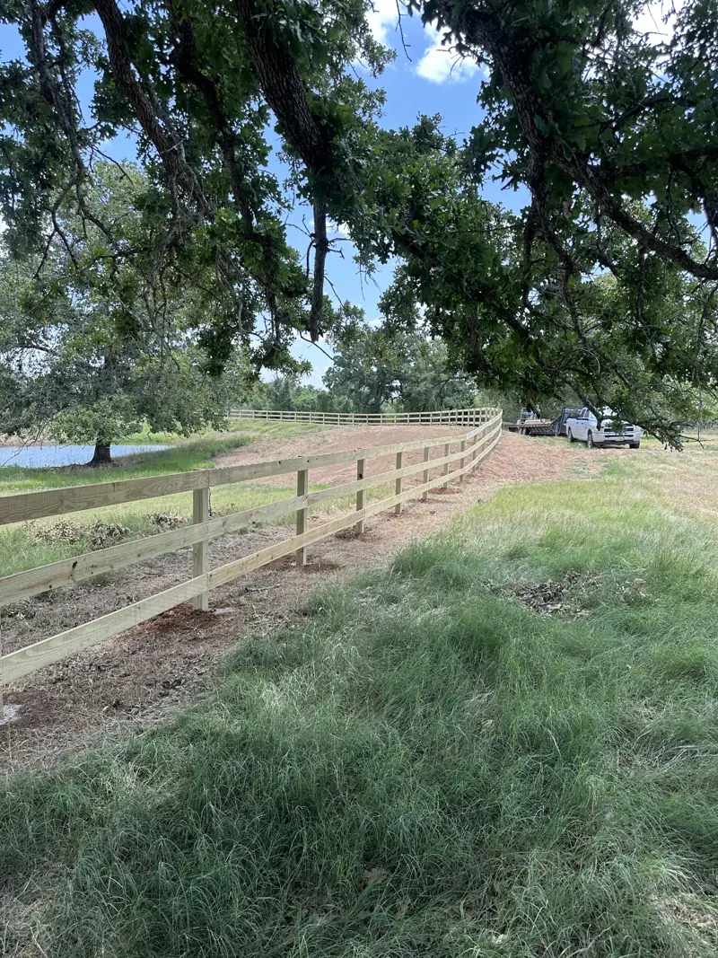 New board fence under oak branches, curving past a pond toward a parked pickup and flatbed trailer.
