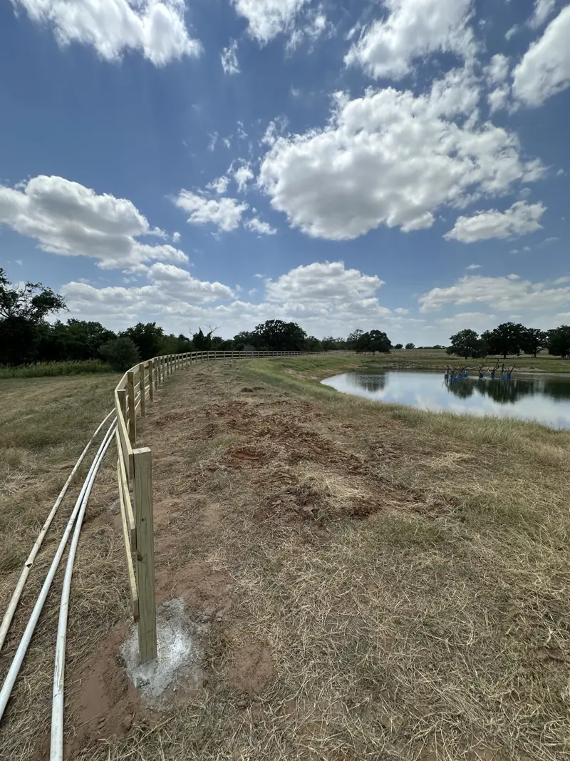 New board fence curving along a pond bank, with a fresh concrete footing at the nearest post.