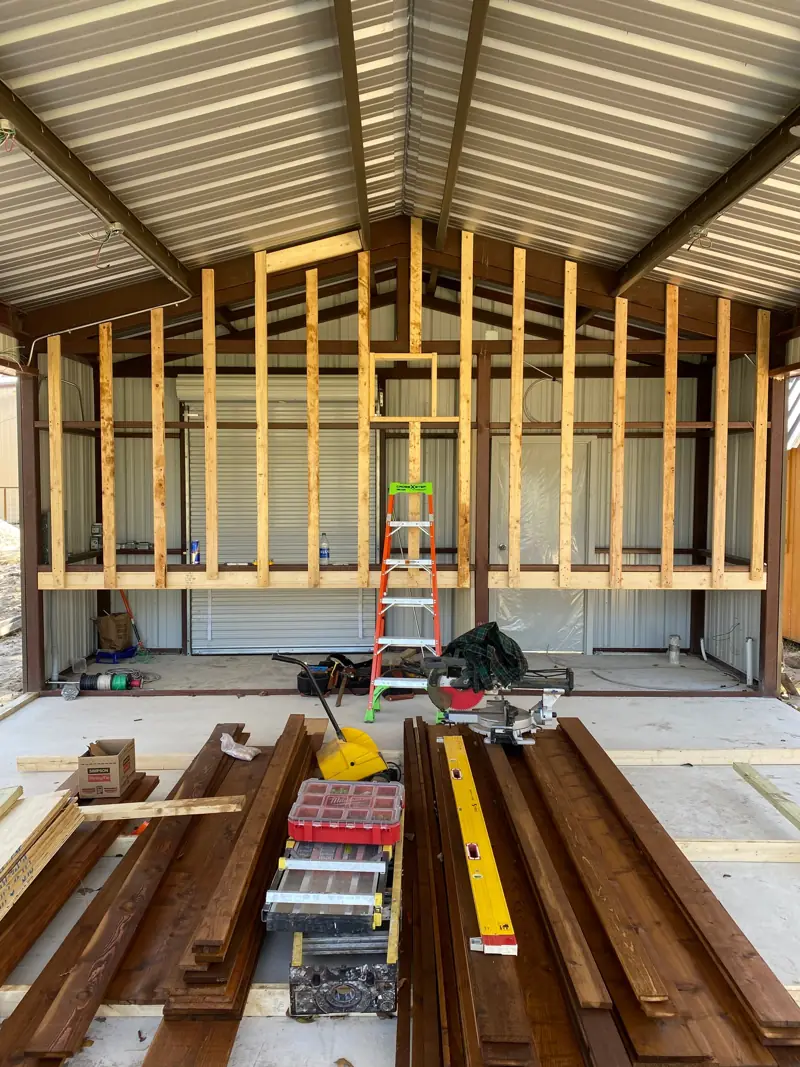 Kitchen wall framing inside a metal pavilion, reclaimed boards on the slab.