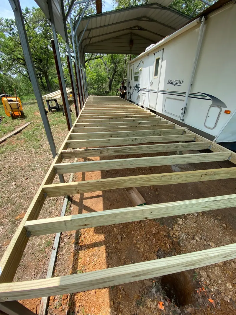 Deck joists beside an RV parked under a metal RV cover.