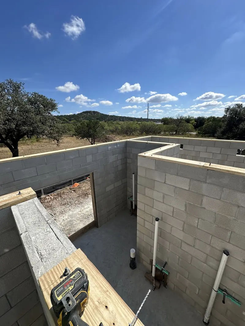 Concrete block walls with wood top plates bolted on, plumbing pipes inside, and wooded hills beyond.
