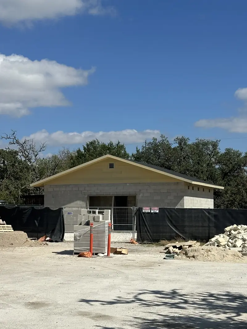 Concrete block building with dark shingle roof and pale gable siding, behind site fencing and block pallets.