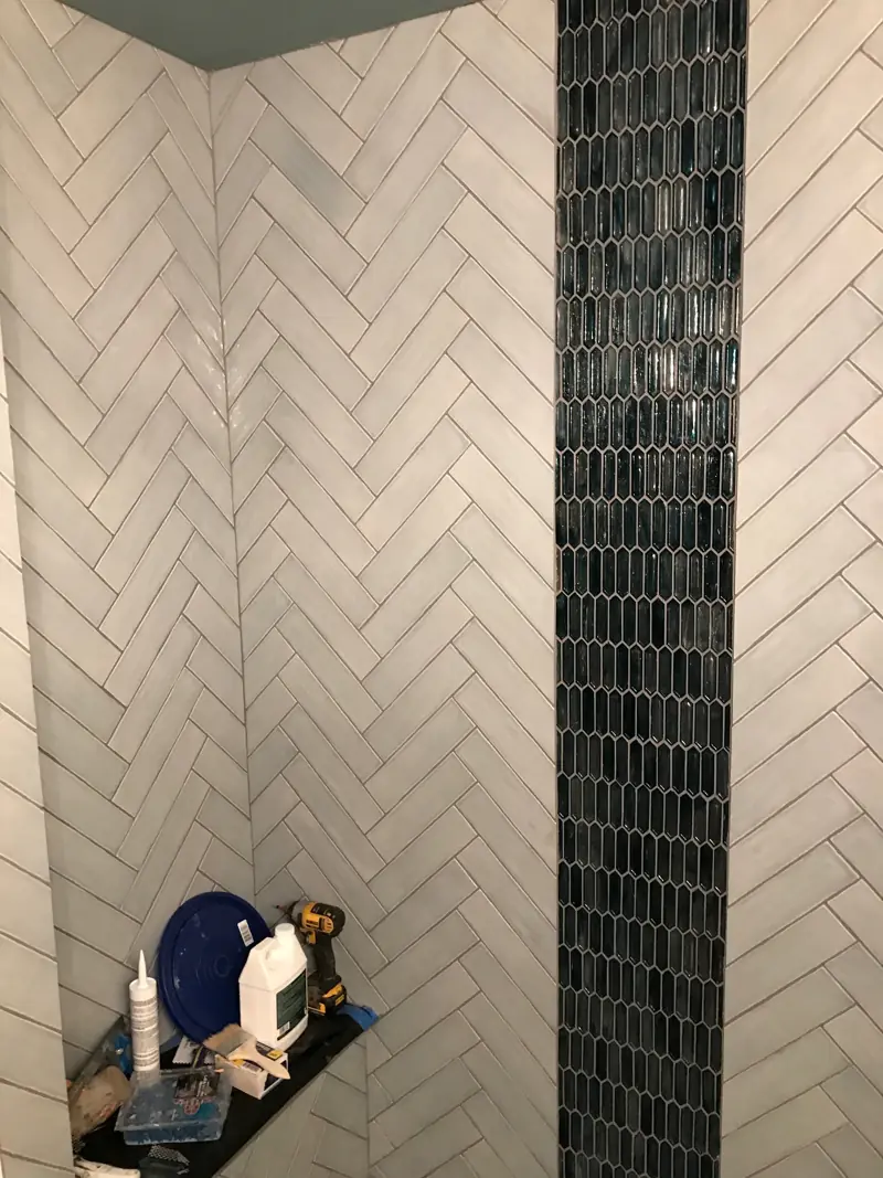 Corner of the shower with herringbone tile, the blue mosaic stripe and a corner shelf.