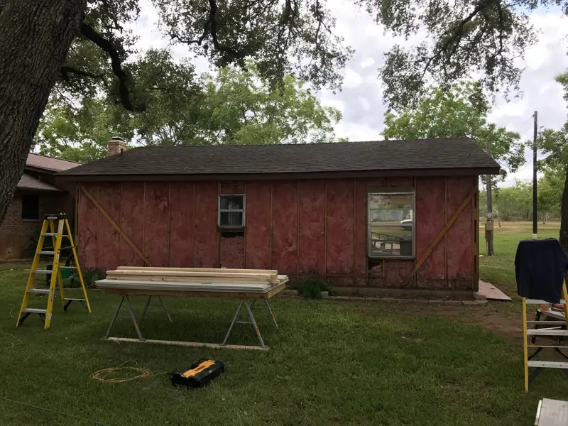 Back wall of a ranch house with the old siding off, down to the red sheathing, lumber on sawhorses.